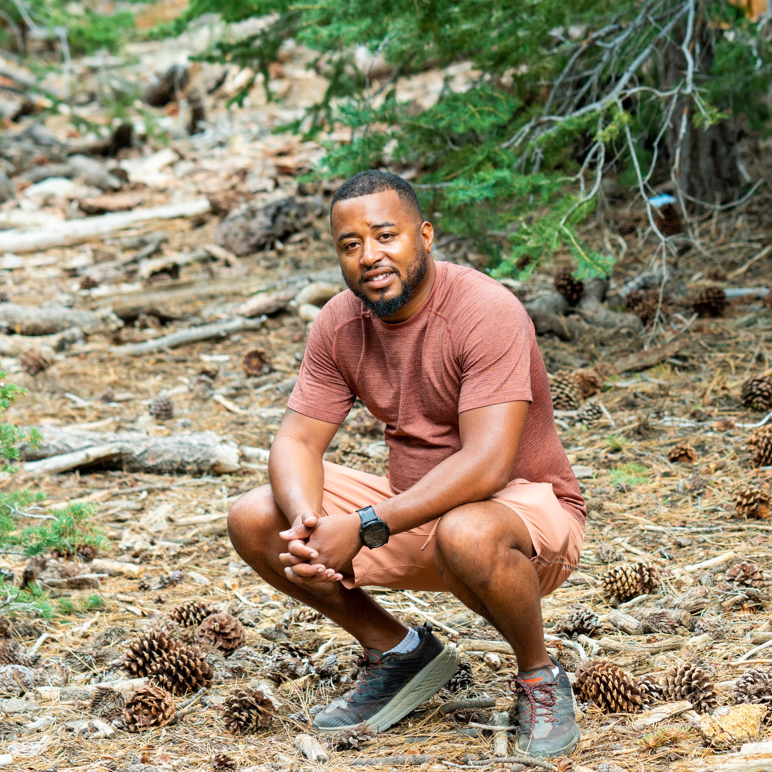 A man crouching in the woods with pine cones, telling an interesting story About Us.