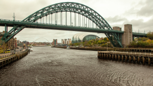 The photo shows a large green arch bridge spanning a wide river, with a cityscape and modern buildings visible in the background on a cloudy day, reminiscent of the scenic views along the Great North Run route.