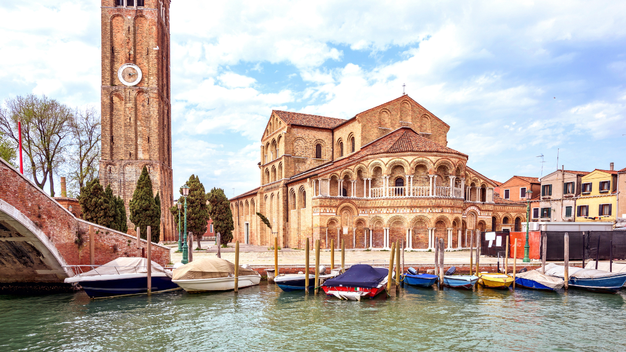 A historic brick church with a tall bell tower stands by a canal in Venice, with several boats tied to wooden posts in front, under a partly cloudy sky.