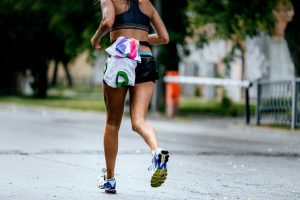 Person running on a wet street, wearing a sports bra, shorts, and running shoes, with a jacket tied around their waist. Trees and a barrier are in the background, evoking the determination needed for running your first race from a 5K to marathon.