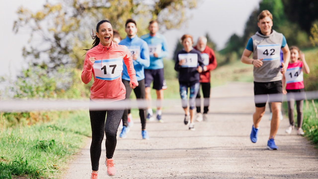 Group of runners on a trail with race bibs. Woman in pink sweatshirt with bib number 17 leading, followed by other runners with bib numbers 42, 6, and more. The race is set against lush greenery in the background.