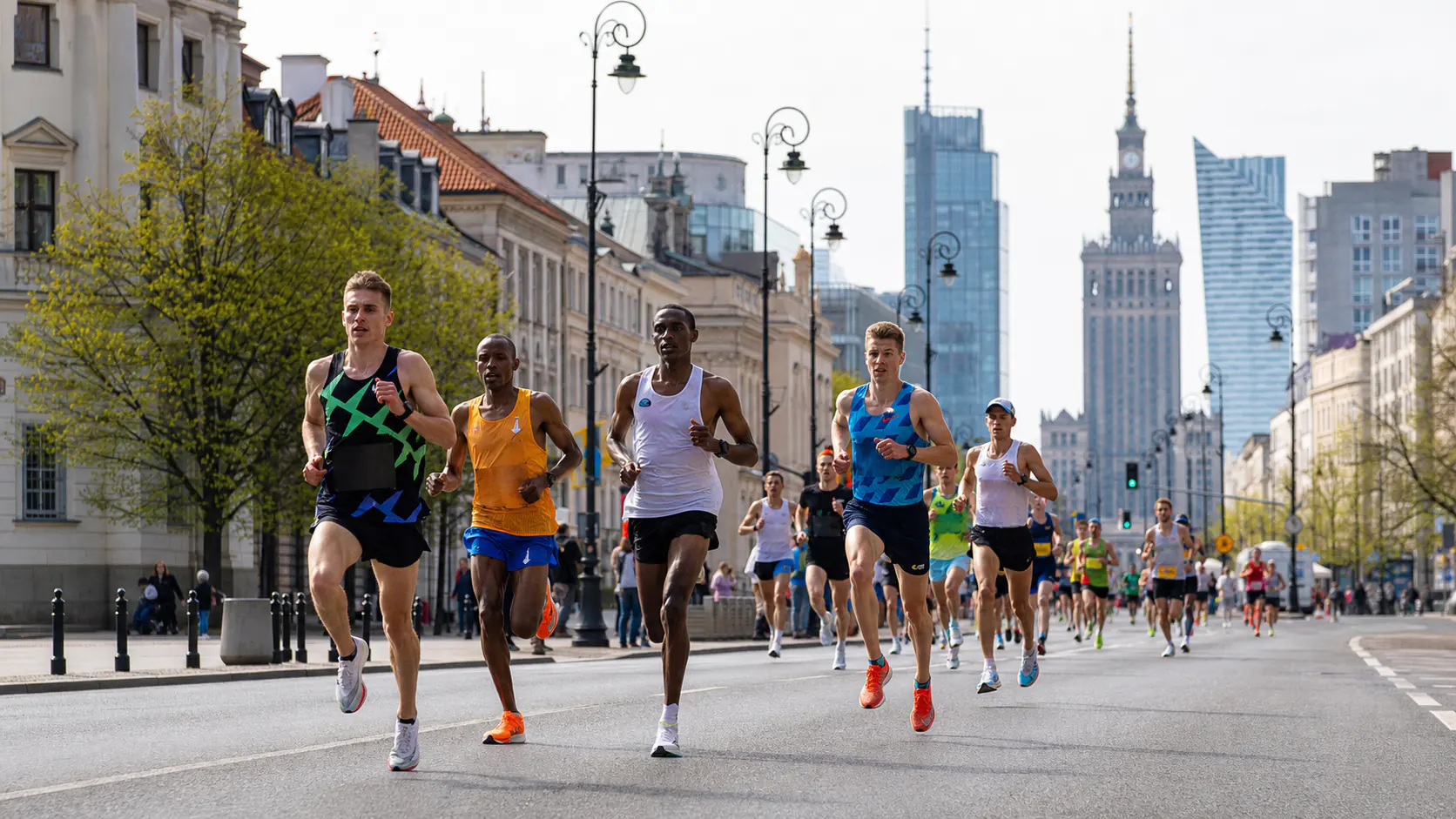 Group of runners sprinting down a wide city street during a race, with historic buildings and modern skyscrapers in the background.